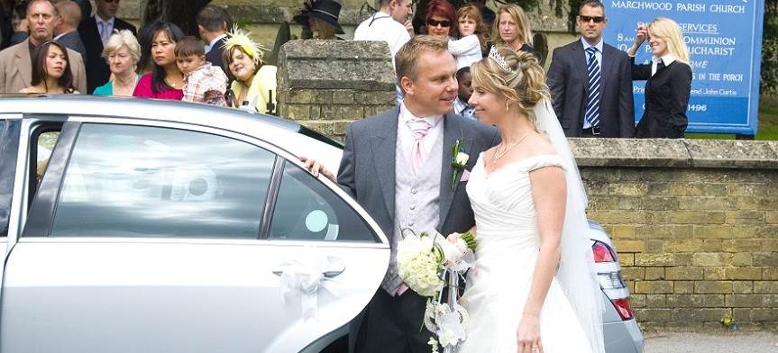 image of a wedding couple standing by a Mercedes Wedding Chauffeur Car with some of the guest standing by
